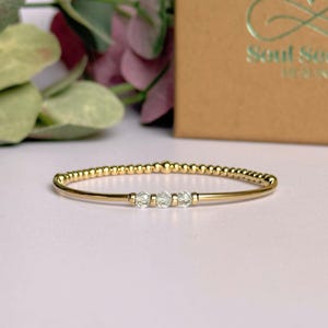 May include: A gold-colored bracelet with three light green, faceted beads. The bracelet has a delicate design with small, round gold beads along the top. The background includes a blurred box with the text "Soul Seek Healing".