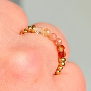 May include: A beaded ring featuring a gold band and a row of faceted, colourful gemstones. The gemstones transition from clear to orange and red. The ring is shown on a person's finger.