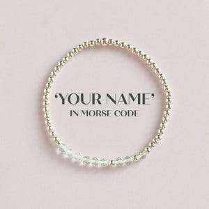 May include: A silver-coloured beaded bracelet with clear crystal beads. The text "YOUR NAME" IN MORSE CODE is displayed on a soft pink background. The bracelet is circular in shape.