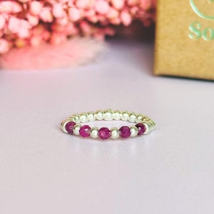 May include: A silver ring featuring a beaded band and a line of bright, round, ruby-coloured gemstones. The ring is set against a soft pink backdrop, with a blurred floral arrangement and a gold box in the background.