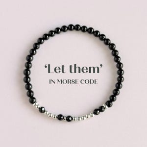 Könnte beinhalten: Schwarzes Perlenarmband mit silbernen Akzenten. Das Armband zeigt den Satz 'Let them' in Morsecode. Die Perlen sind klein und rund, zu einem Kreis aufgereiht. Der Hintergrund ist ein zartes Rosa.
