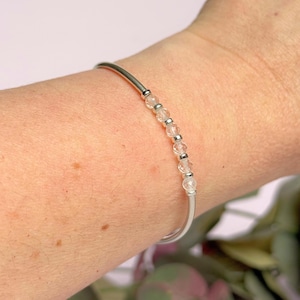 May include: A silver bangle bracelet with clear, faceted beads. The bracelet is worn on a wrist, highlighting the delicate design and the sparkle of the beads. The bracelet is a simple, elegant piece of jewellery.