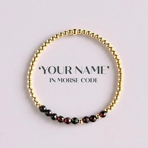 May include: Gold beaded bracelet with a section of dark red beads. The bracelet has the text "YOUR NAME" in Morse code. The bracelet is on a white background.