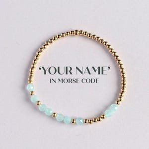 Könnte beinhalten: Goldperlenarmband mit hellblauen Edelsteinakzenten. Das Armband trägt den Text "YOUR NAME" in Morsecode. Das Armband befindet sich auf weißem Hintergrund.