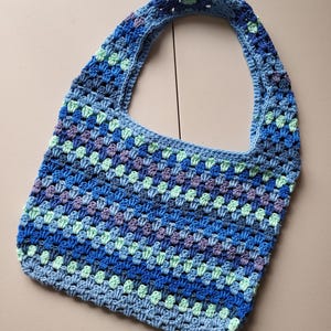 Pu&ograve; includere: Una borsa tote all'uncinetto fatta a mano nei toni del blu, verde e viola. La borsa presenta un motivo a granny square e una singola tracolla. La borsa &egrave; realizzata in filato morbido ed &egrave; perfetta per trasportare oggetti di uso quotidiano.