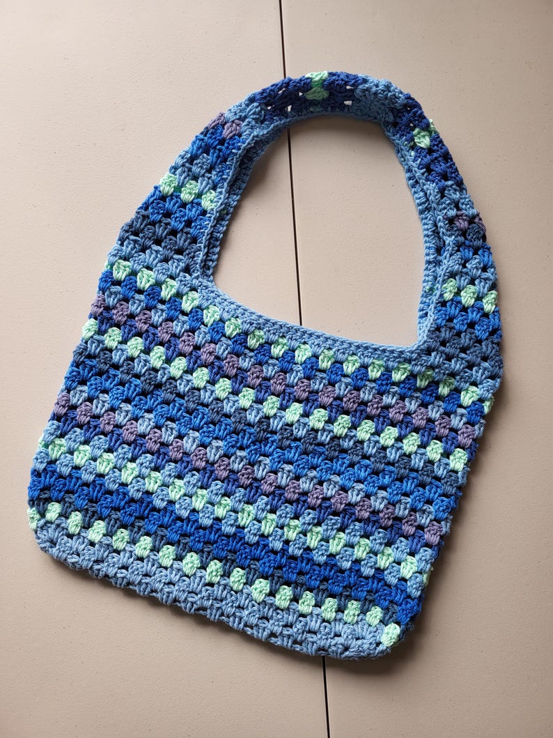Pu&ograve; includere: Una borsa tote all'uncinetto fatta a mano nei toni del blu, verde e viola. La borsa presenta un motivo a granny square e una singola tracolla. La borsa &egrave; realizzata in filato morbido ed &egrave; perfetta per trasportare oggetti di uso quotidiano.