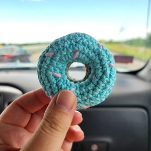 Peut inclure: Un donut crocheté avec un glaçage bleu clair et des vermicelles roses. Le donut a un centre marron et est tenu devant un arrière-plan flou d'un intérieur de voiture et d'un ciel clair.