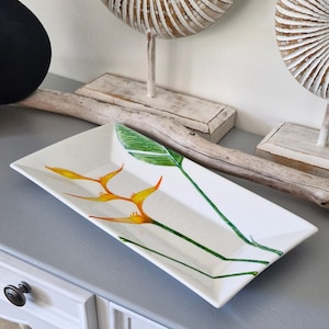May include: Rectangular white ceramic serving tray with a hand-painted floral design. The design features orange and yellow bird of paradise flowers with green stems and leaves. The tray is displayed on a grey surface.
