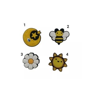 May include: Four colourful shoe charms. The charms include a yellow moon with a star, a bee with black and yellow stripes, a white daisy with a yellow centre, and a smiling yellow sun with black accents. These charms are designed to decorate shoes.