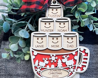 May include: A wooden Christmas ornament shaped like a mug with marshmallows stacked on top. The marshmallows are decorated with smiley faces and have names written on them: Dustin, Hunter, Noah, Lanie, Mom, and Dad. The mug is red with a white snowflake pattern and a reindeer design. The ornament has a red and black plaid ribbon attached to it. The text "Schaule Christmas 2022" is written on the bottom of the mug.
