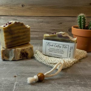 May include: Two bars of handmade soap with a layered, textured appearance, one stacked on top of the other. The soap is light brown with darker brown stripes and topped with dried flowers. A small terracotta pot with a cactus and a soap saver bag are also in the image. The soap is labeled "Black Cactus Tide".