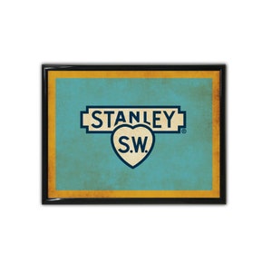 May include: A vintage-style framed print with a blue and yellow color scheme. The print features the text "STANLEY S.W." in a white heart shape with a blue outline. The text is surrounded by a blue and yellow banner with the text "STANLEY" in white.
