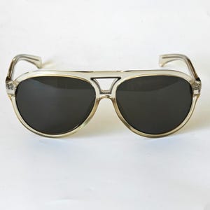 May include: Aviator sunglasses featuring a translucent, light beige plastic frame with a double bridge design and dark, solid black lenses. The temples are curved at the ends, and the overall construction provides a classic, full-rimmed look.