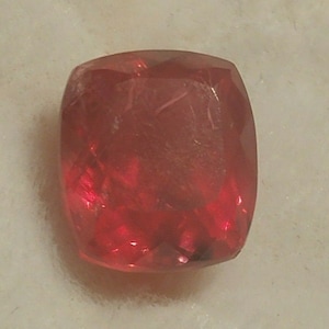 Può includere: Una pietra preziosa di colore rosso intenso con taglio sfaccettato. La pietra preziosa ha una forma quadrata con angoli arrotondati.