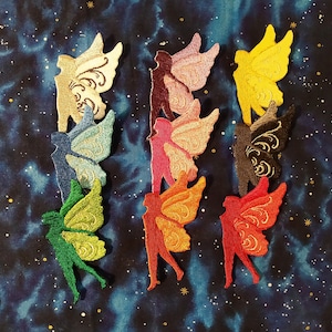 May include: A collection of embroidered fairy patches in various colors, including green, blue, yellow, and red. Each patch features a fairy silhouette with detailed wing designs. The patches are arranged on a dark blue fabric with a starry pattern.
