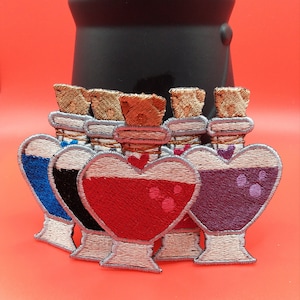 May include: Five heart-shaped glass bottles with cork stoppers and different colored liquids inside. The bottles are arranged in a row, with the red bottle in the center. The bottles are embroidered with a silver thread.