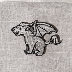 May include: A gray embroidered patch of a small, winged creature with a long tail. The creature has a pointed snout and large, rounded ears. The wings are spread out and have a black outline.