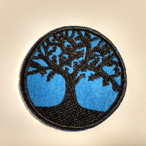 May include: A blue and black embroidered patch featuring a tree of life design. The tree is black and the background is blue.