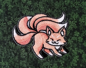 May include: Embroidered patch of a cartoon fox with nine tails. The fox is orange and white with a smiling face. The tails are curled and layered, creating a swirling pattern.