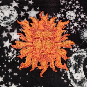 May include: Embroidered orange sun patch with a detailed face and flame-like rays. The patch is set against a black background with white stars and celestial bodies, creating a cosmic theme. The sun's face has a stern expression.
