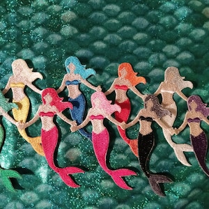 May include: A collection of embroidered mermaid patches in various colors, including green, yellow, pink, red, blue, and purple. Each mermaid is holding hands, set against a textured, teal-green background.