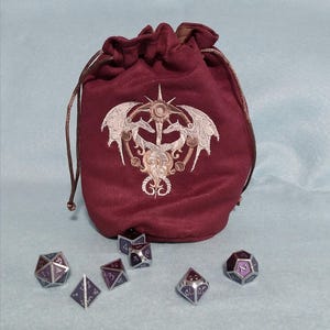 May include: A burgundy velvet dice bag with a drawstring closure and an embroidered design featuring two dragons and a sunburst. Six metal dice with purple and silver accents are scattered around the bag. The bag is designed for tabletop gaming.