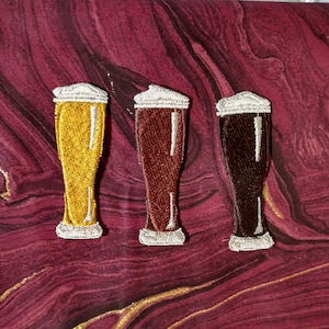 May include: Embroidered patches featuring three beer glasses with foamy heads. The glasses are filled with golden, amber, and dark brown beer. The patches have a white border and are set against a burgundy and gold marbled background.