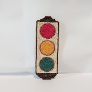 May include: A brown and white embroidered patch of a traffic light with red, yellow, and green lights.