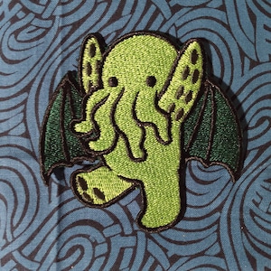 May include: Embroidered patch featuring a green Cthulhu design with black outlines and dark green wings. The patch is on a blue fabric background with a swirling pattern. The Cthulhu figure has tentacles and is in a walking pose.