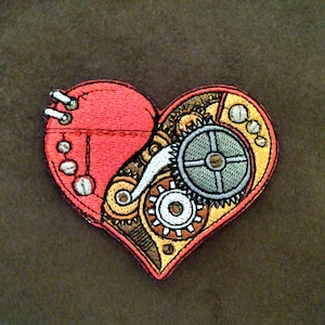 May include: A red heart-shaped patch with a detailed embroidered design of gears and cogs inside. The heart is outlined in yellow and has a stitched appearance.