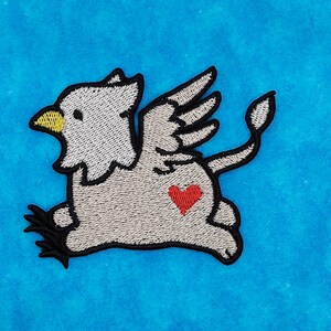 May include: A gray embroidered patch of a winged creature with a heart on its chest. The creature has a bird-like head and a lion-like body with a tail.