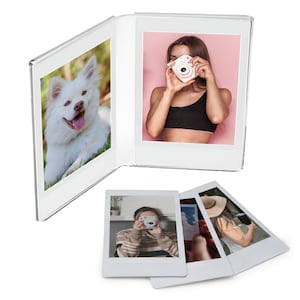 May include: A white photo album with a clear cover, open to display two photos. One photo features a white dog, the other a person holding a white camera. Several instant photos are scattered below.