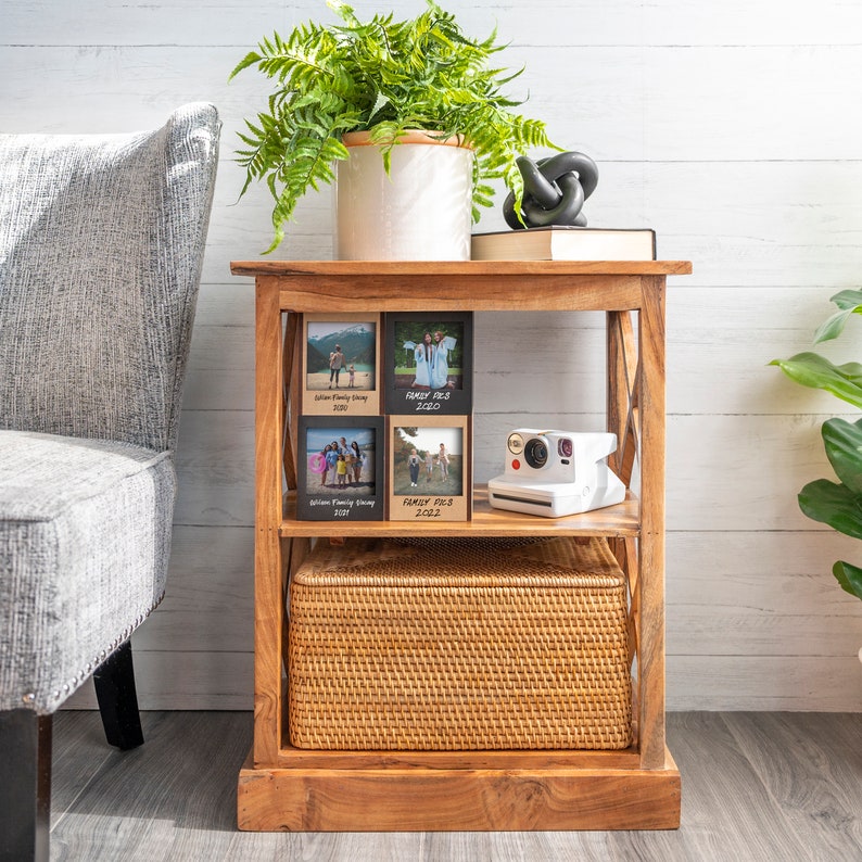 Op de afbeelding: Een houten bijzettafel met een geweven mand op de onderste plank en een witte instant camera op de middelste plank. De tafel toont ingelijste foto's met het label "Family Pics" uit 2020, 2021 en 2022. Een potvaren en een decoratieve knoop staan bovenop.