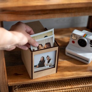 Op de afbeelding: Een houten fotodoos met een rechthoekig venster, met daarin verschillende afgedrukte foto's. Een hand plaatst een foto in de doos. Een witte Polaroid-camera met een regenboogstreep staat op de plank.