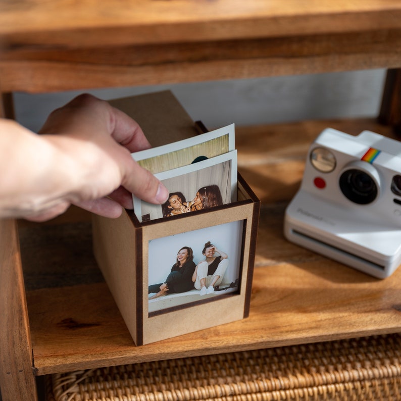Op de afbeelding: Een houten fotodoos met een rechthoekig venster, met daarin verschillende afgedrukte foto's. Een hand plaatst een foto in de doos. Een witte Polaroid-camera met een regenboogstreep staat op de plank.