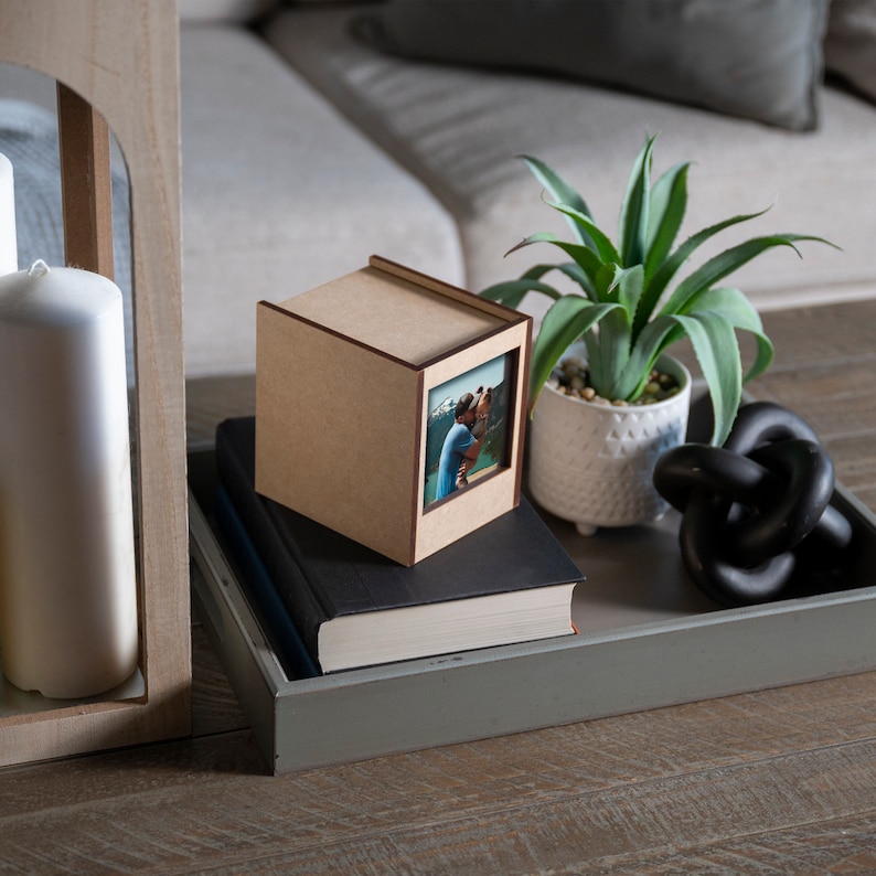 Op de afbeelding: Een kleine, lichtbruine houten fotodoos met een zichtbare foto aan &eacute;&eacute;n kant, rustend op een stapel boeken en een grijze dienblad. Een groene plant in een pot en een zwarte decoratieve knoop staan ook op de dienblad.