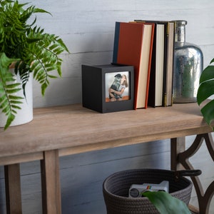 Op de afbeelding: Een houten console met decoratieve items. Een zwart fotolijstje, boeken en een zilveren glazen fles zijn op de tafel geplaatst. Een varen en een plant met grote bladeren voegen groen toe. Een geweven mand met een speelgoedcamera staat eronder.