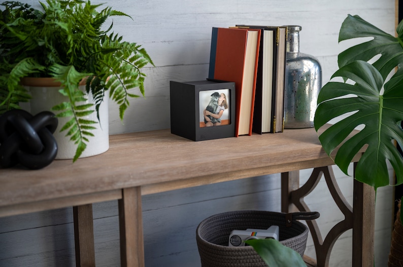Op de afbeelding: Een houten console met decoratieve items. Een zwart fotolijstje, boeken en een zilveren glazen fles zijn op de tafel geplaatst. Een varen en een plant met grote bladeren voegen groen toe. Een geweven mand met een speelgoedcamera staat eronder.