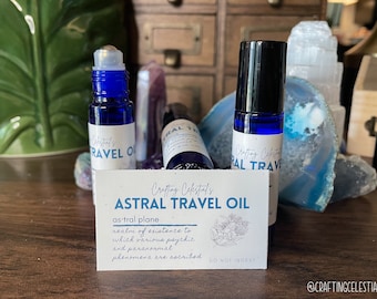 May include: Three blue glass roll-on bottles labeled "Crafting Celestial's ASTRAL TRAVEL OIL" are displayed with a card that reads "astral plane realm of existence". The bottles are surrounded by crystals and other decorative items.