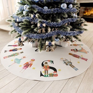 May include: A white Christmas tree skirt with a colorful Nutcracker and ballerina design. The skirt features the letter "S" and is decorated with stars. A decorated Christmas tree with blue ornaments and garland sits above the skirt.