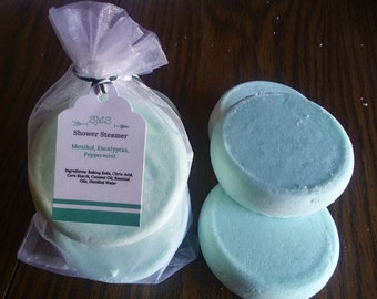 May include: A set of three round, light blue shower steamers in a white organza bag with a tag that reads "Shower Steamer, Menthol, Eucalyptus, Peppermint. Ingredients: Baking Soda, Citric Acid, Corn Starch, Coconut Oil, Essential Oils, Distilled Water."