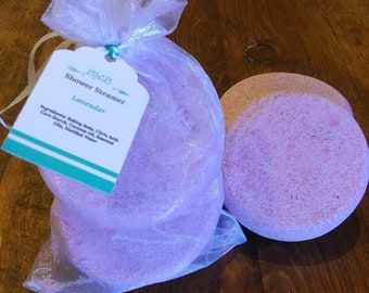 May include: Two lavender-colored shower steamers in a white mesh bag. The bag has a tag that reads "SMS Shower Steamer Lavender" and lists the ingredients: Baking Soda, Citric Acid, Corn Starch, Coconut Oil, Essential Oil, Distilled Water.