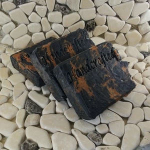 May include: Three bars of black soap with orange swirls. The soap is handmade and has the word "Handcrafted" stamped on each bar.