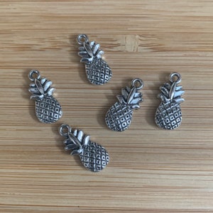 May include: Five silver pineapple charms with a textured surface. The charms are on a wooden background.
