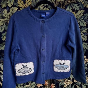 May include: A navy blue cardigan with two square pockets, each featuring a moth applique. The cardigan has a round neckline and a button-up front. The moth appliques are in shades of gray and blue.