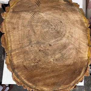 Puede incluir: Una sección transversal de un tronco de árbol, que muestra la veta natural de la madera y los anillos de crecimiento. La madera tiene un tono marrón cálido con áreas más oscuras y claras, y un borde de corteza rugoso y texturizado. La forma circular resalta la edad y la historia del árbol.
