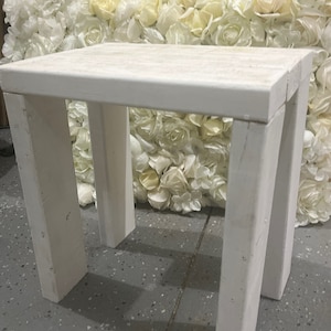 Puede incluir: Pequeña mesa de madera blanca con tapa rectangular y cuatro patas robustas. La mesa está pintada de blanco y tiene un aspecto rústico. El fondo presenta una pared de flores artificiales blancas y crema.