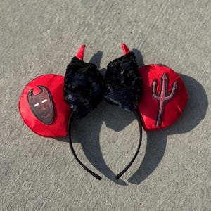 May include: Devil-themed headband with red ears, black sequined bow, and red horns. One ear features a mask graphic, the other a trident. The headband is black.