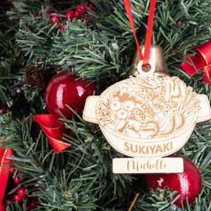 May include: A wooden Christmas ornament shaped like a pot of sukiyaki, a Japanese hot pot dish. The ornament is decorated with a drawing of various ingredients, including vegetables, meat, and noodles. The ornament also has a personalized name tag that reads "Sukiyaki Michelle".