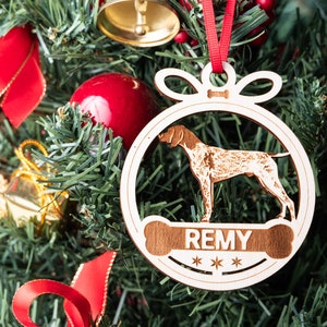 May include: A wooden Christmas ornament shaped like a circle with a dog silhouette inside. The ornament has a red ribbon and a bone-shaped tag with the name "REMY" and three stars.